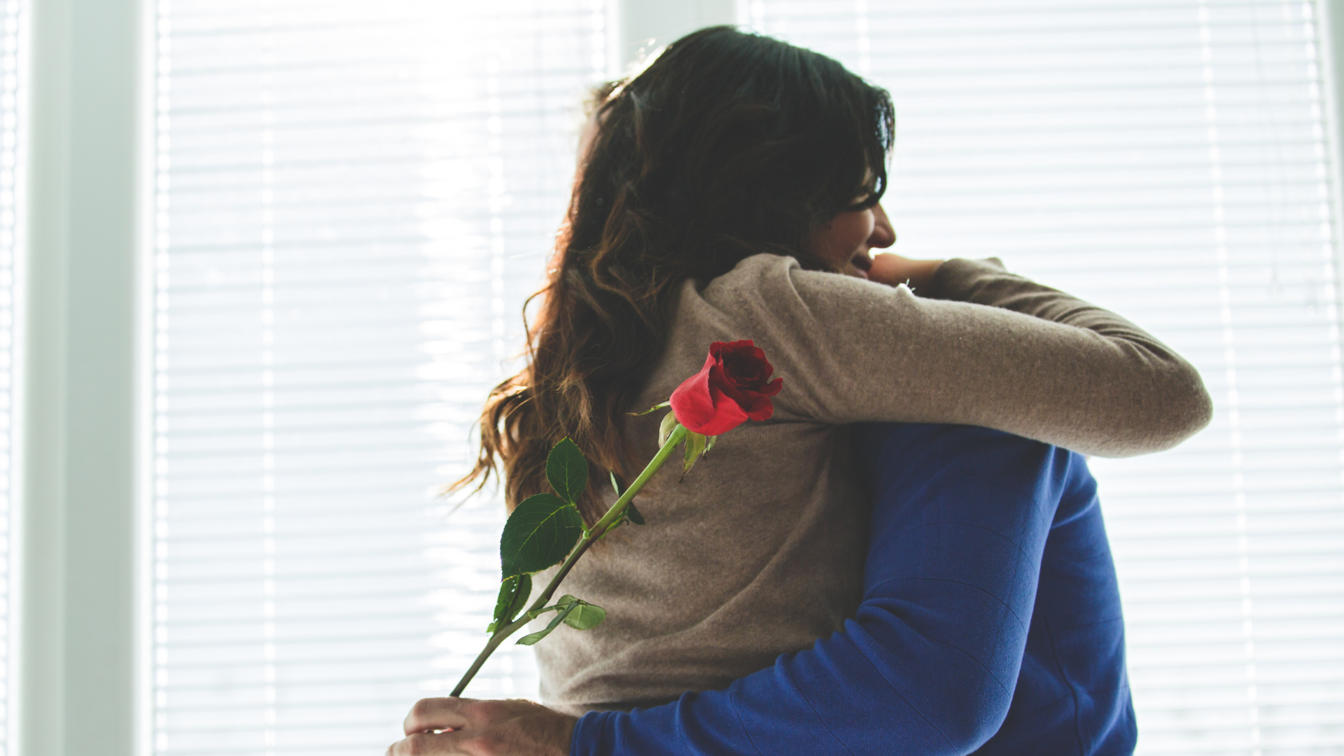 A man is hugging a woman who is holding a red rose behind her back.