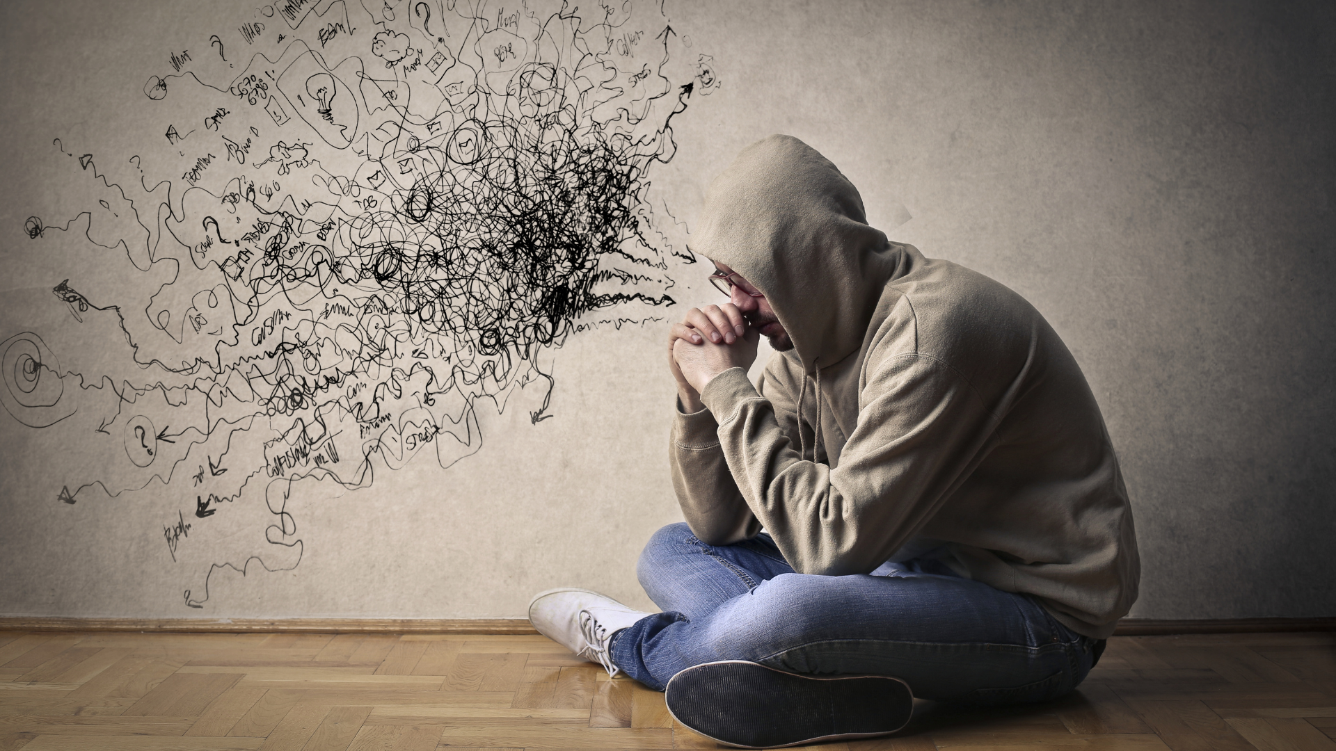 A man in a hoodie is sitting on the floor in front of a wall.