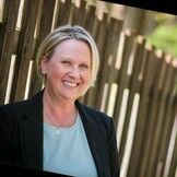 A woman in a suit is smiling in front of a wooden fence.