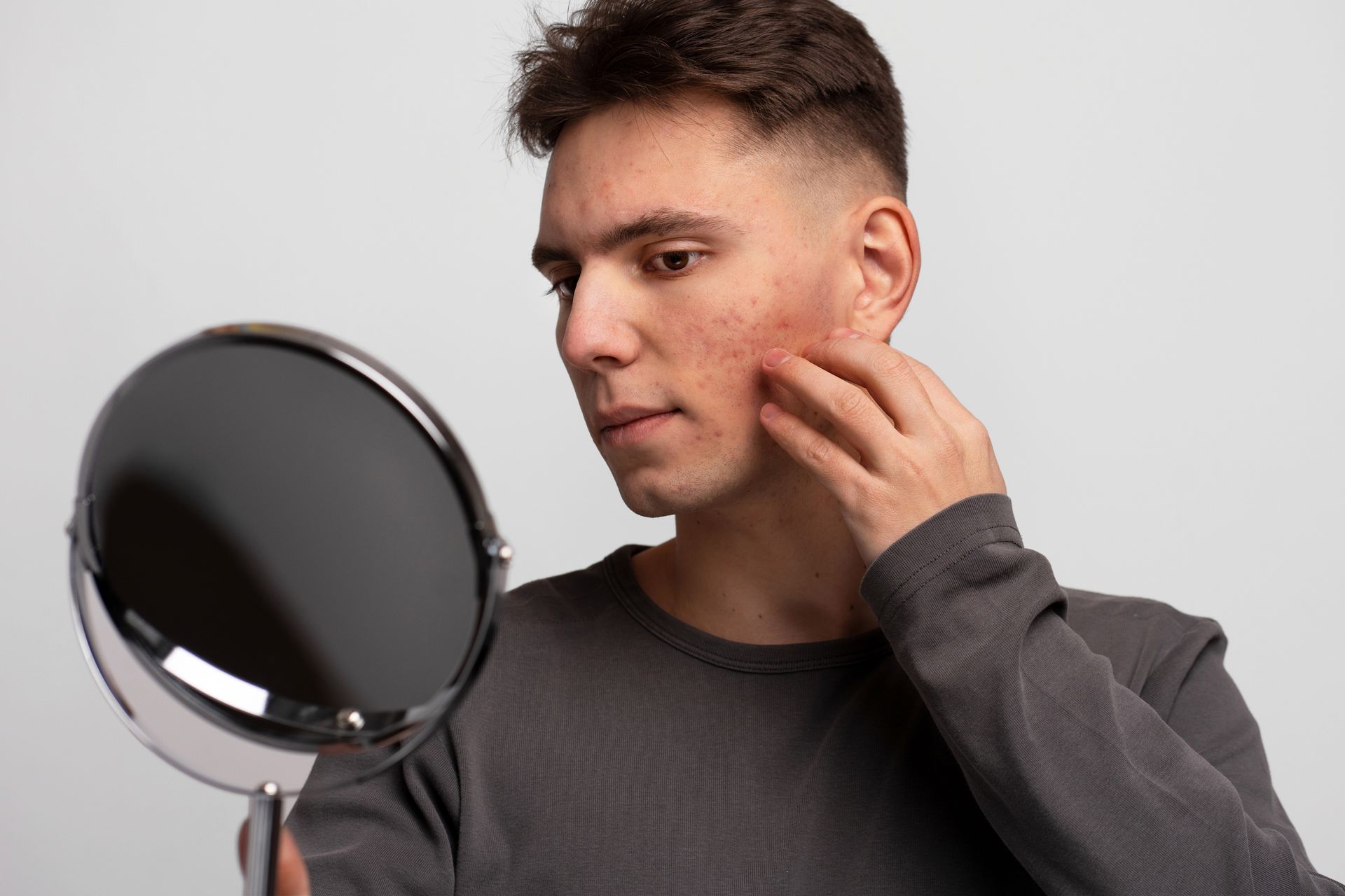 Person examining face in a handheld mirror, touching cheek in a gray shirt on a neutral background