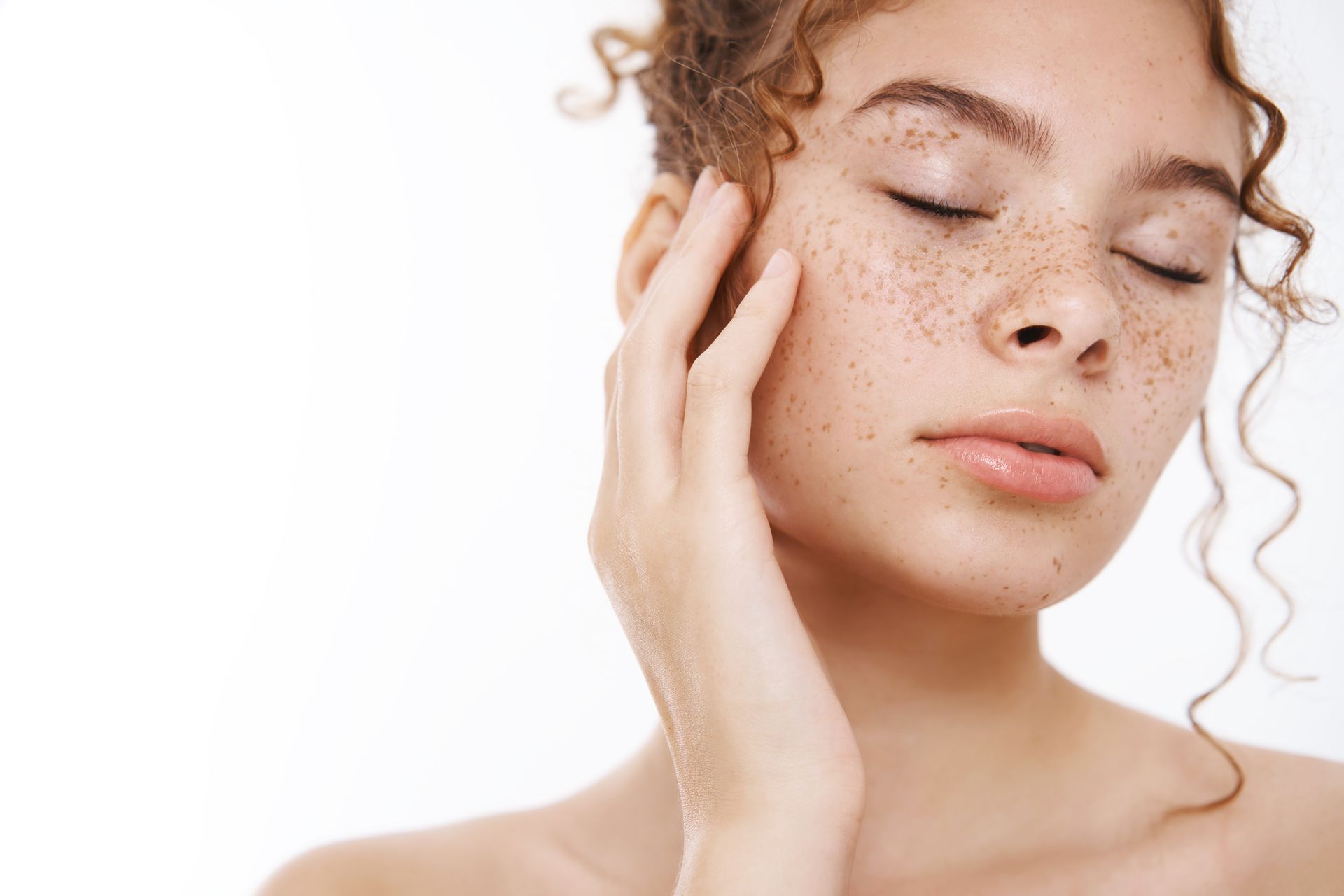 Close-up of a freckled person with eyes closed, gently touching their face against a white background