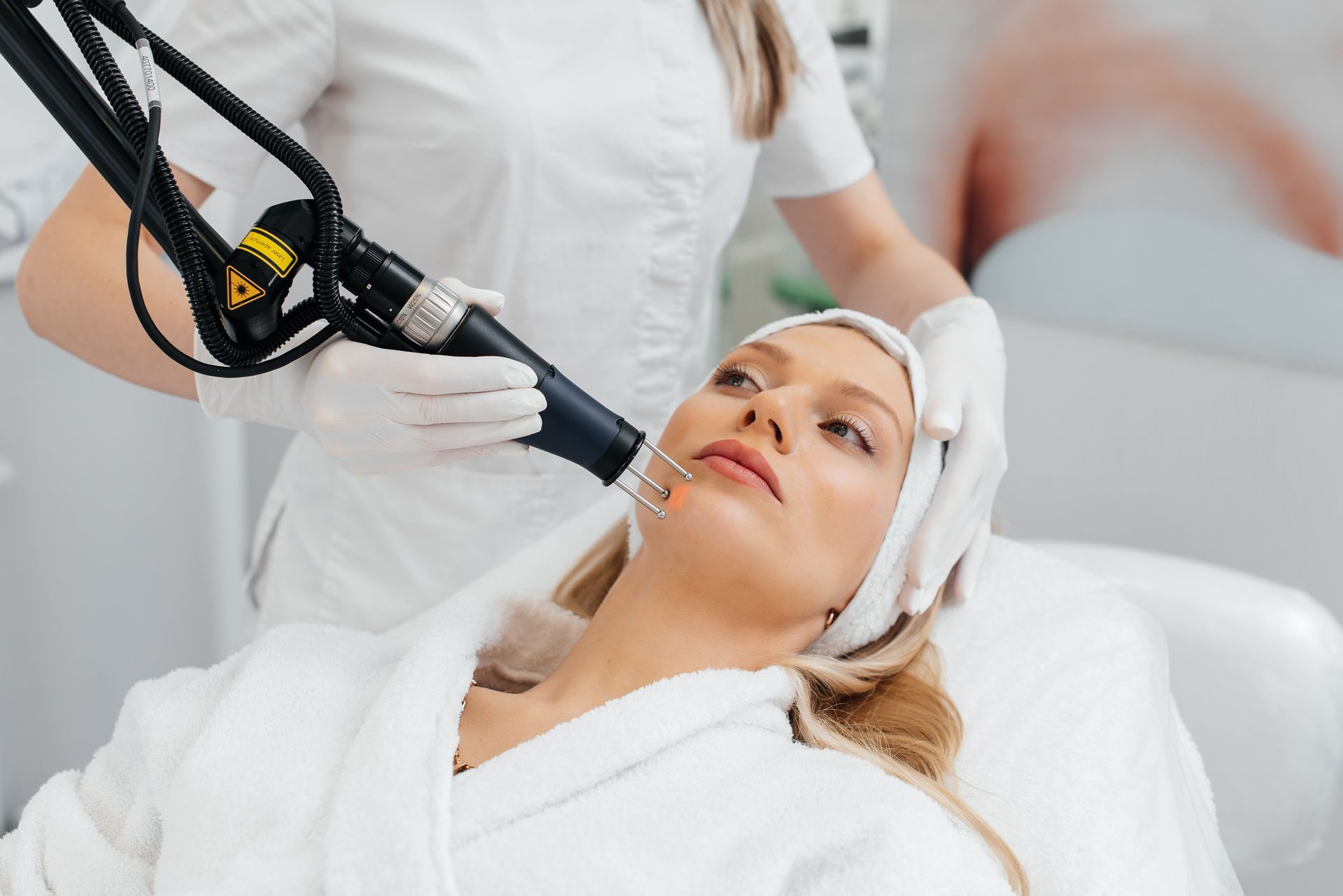Woman receiving facial laser treatment in a spa, with a technician holding the device near her face