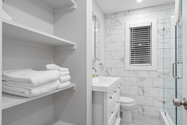 Small white bathroom with shelves of towels, a sink, shower, and window blind