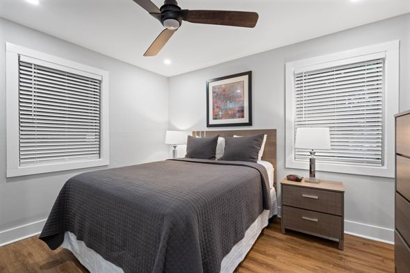 Bright bedroom with a gray bedspread, white walls, two windows, and a ceiling fan.