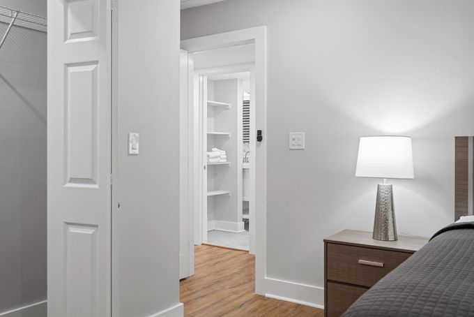 White bedroom with open doorway to a closet, wood floor, nightstand lamp, and bed edge on the right.