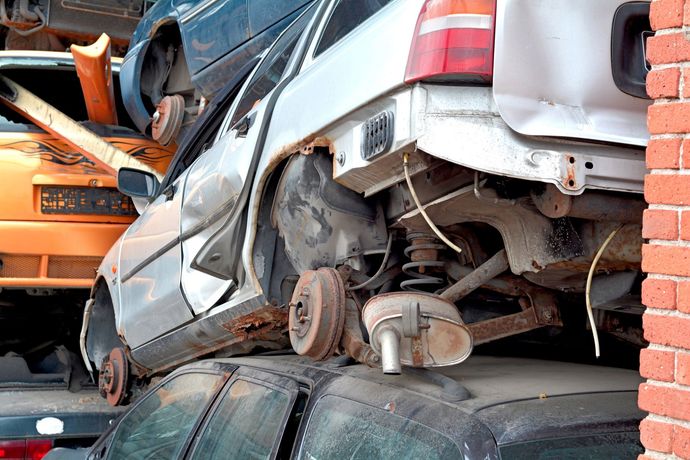 Una carrozzeria argentata, priva di ruote e parti, è accatastata sopra altri veicoli in una discarica, accanto a un muro di mattoni.