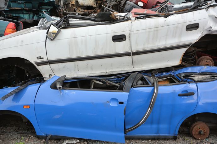 Una carrozzeria argentata, priva di ruote e parti, è accatastata sopra altri veicoli in una discarica, accanto a un muro di mattoni.