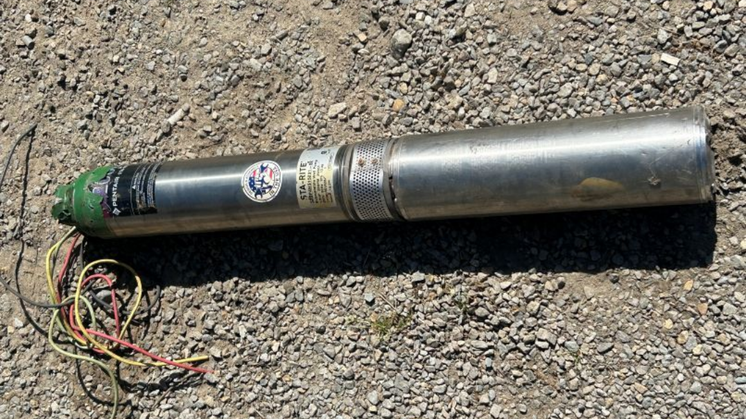 A stainless-steel submersible well pump with colorful power wires attached, lying on a gravel surface.