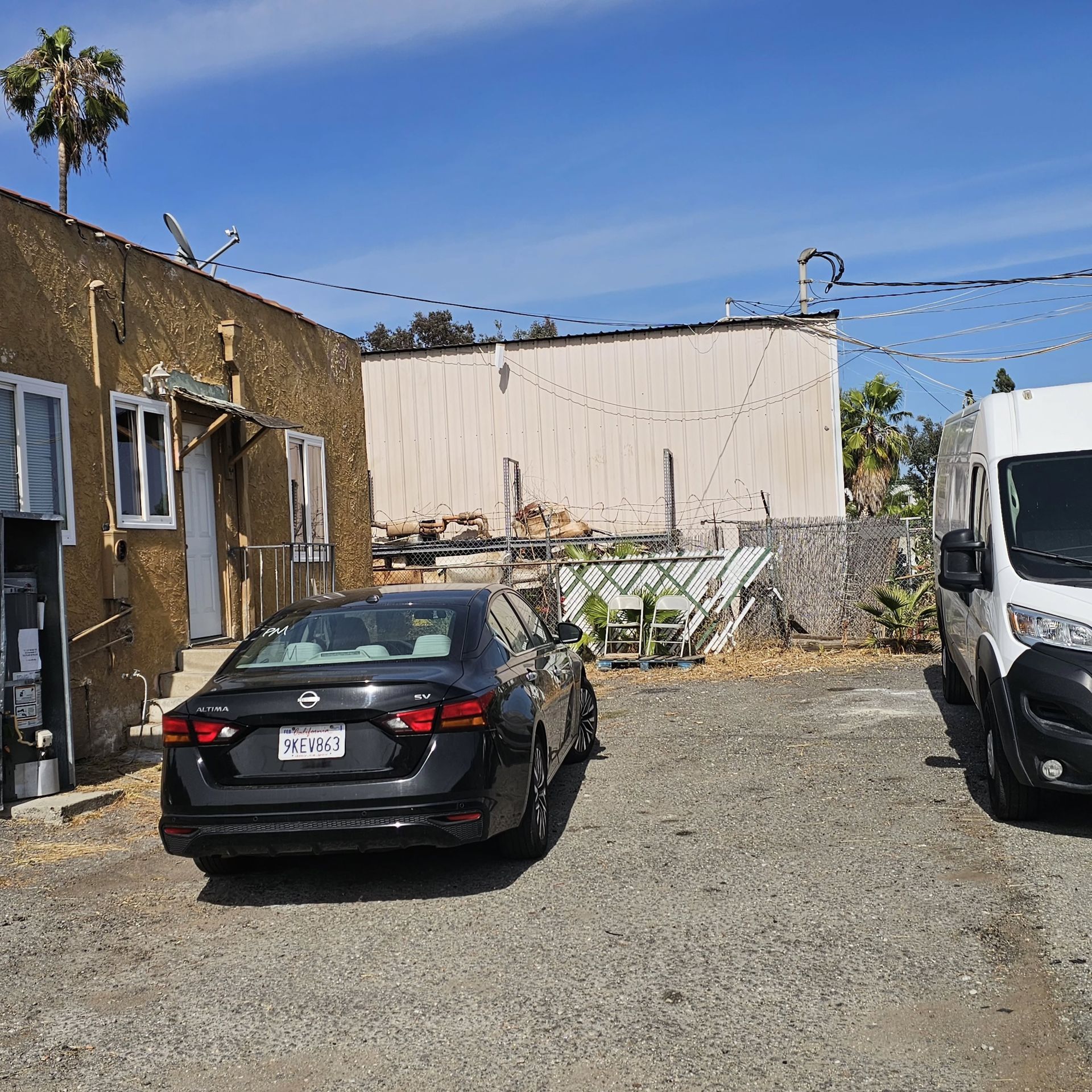 Gravel lot with parked black sedan and white van beside a weathered building under a clear blue sky