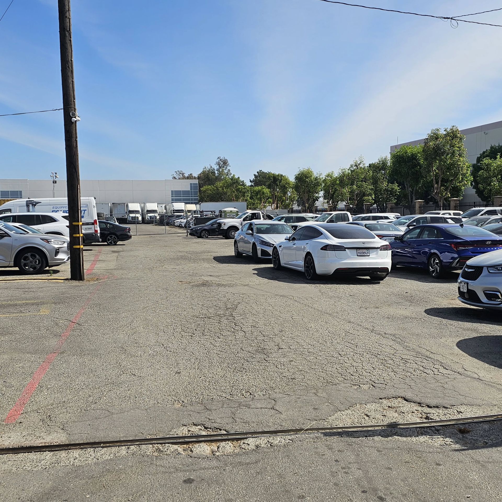 Parking lot filled with parked cars on a sunny day, with a gravel surface and warehouse buildings nearby