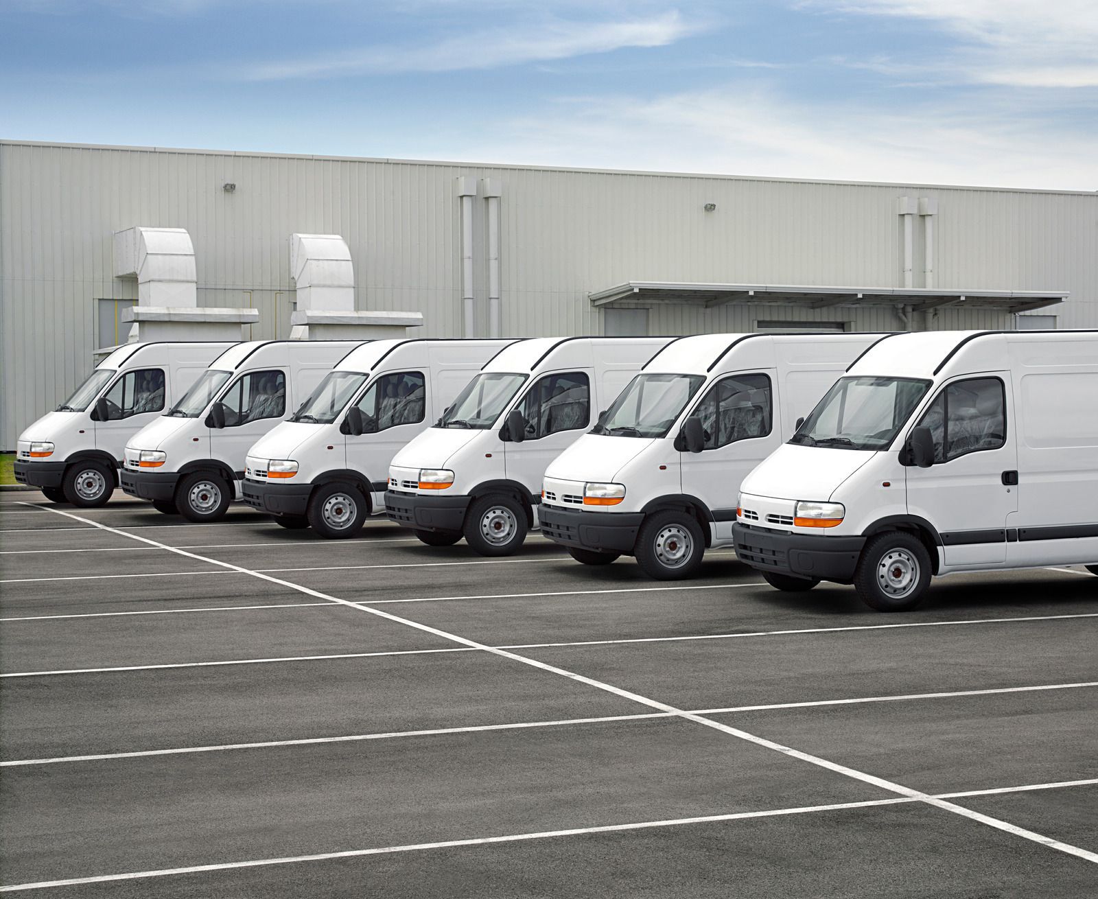 Row of white delivery vans parked in an empty lot beside a warehouse.
