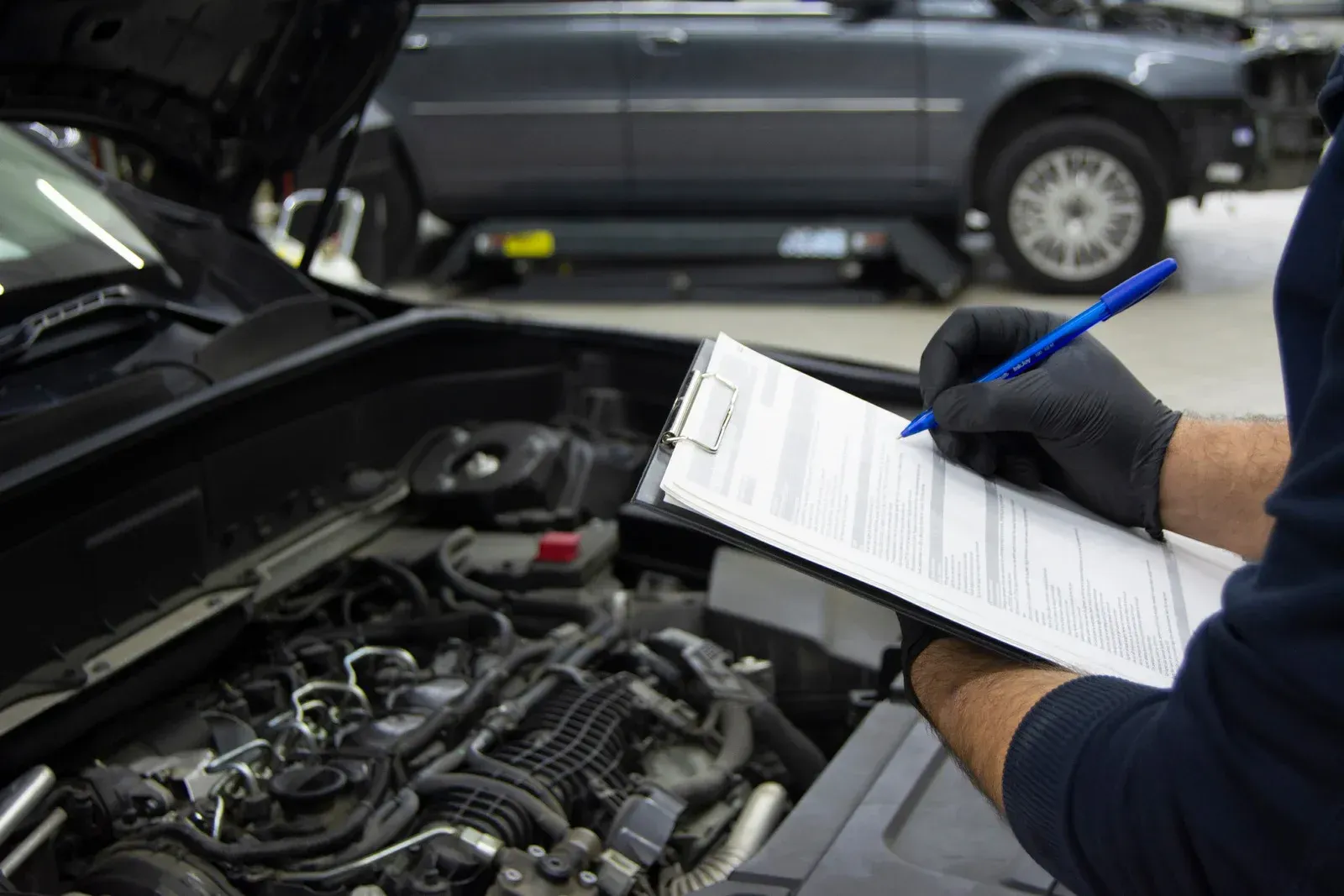 Mechanic in gloves writes on clipboard beside an open car engine in a garage.