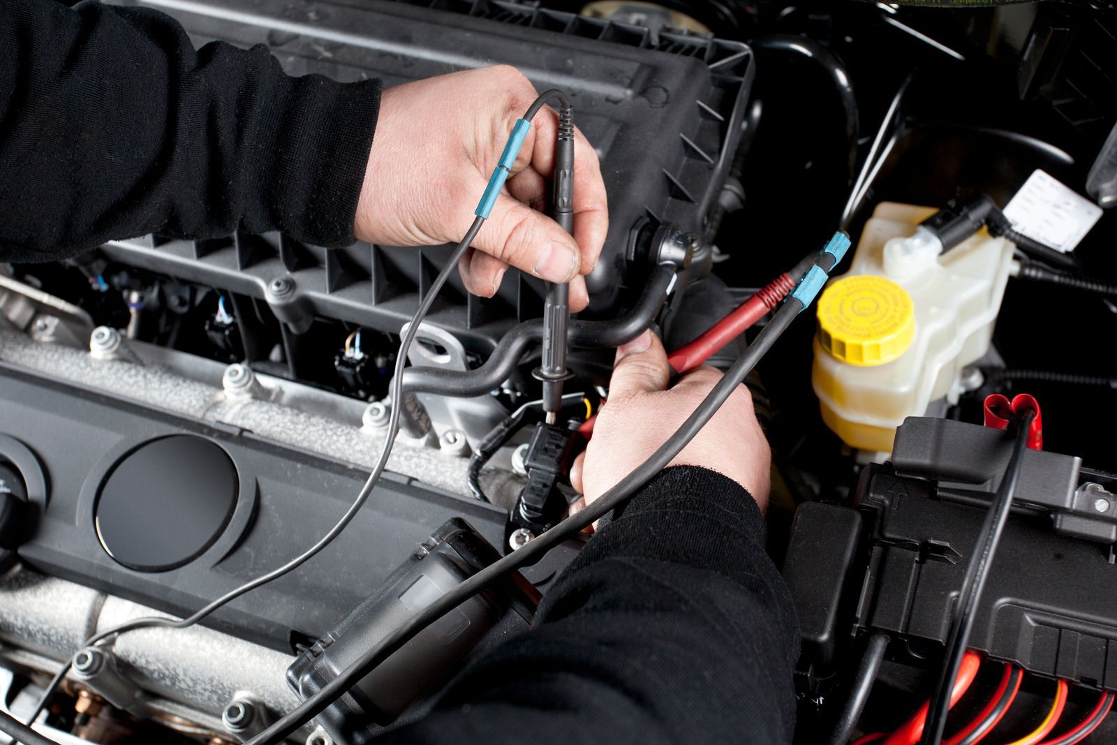 Hands using a multimeter to test wiring in a car engine bay with a yellow fluid reservoir nearby