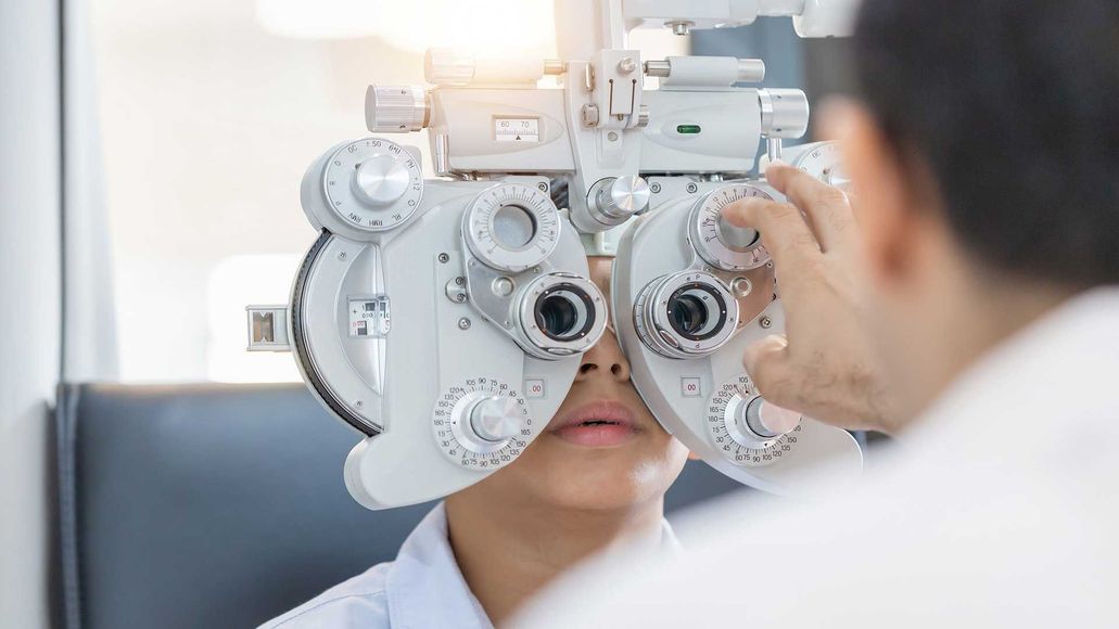Patient viewing an eye exam chart through a phoropter, with an optometrist adjusting the lenses