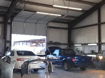 Inside a car repair shop, a blue pickup truck and a white sedan sit near the open garage door. Parts of other vehicles are visible.