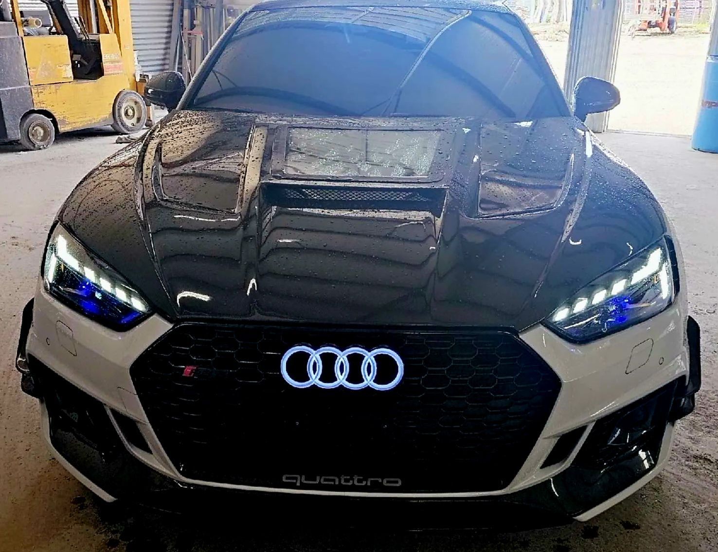 Black Audi Quattro with headlights on, parked indoors with hood raised and front damage visible