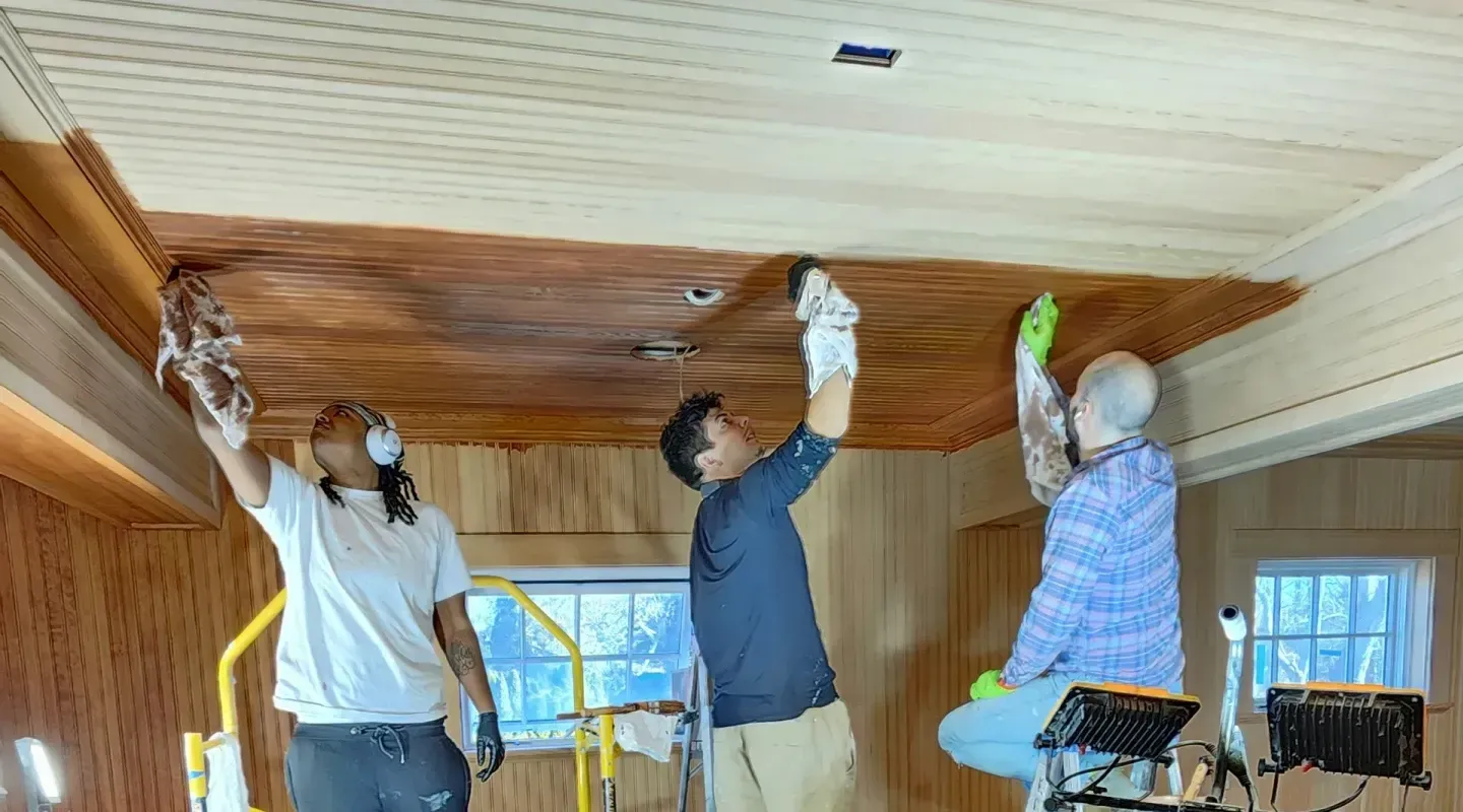 Three people painting a wood-paneled ceiling in a room with large windows.