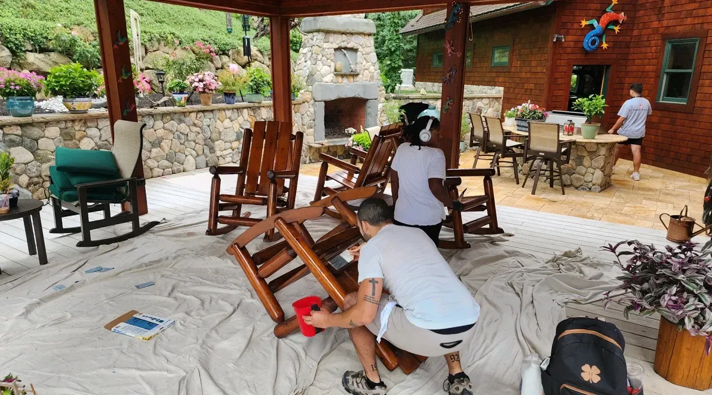 Two people stacking wooden chairs on a patio beside a brick wall and stone fireplace