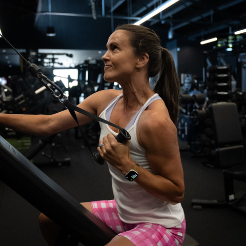 Woman in a gym doing a cable row exercise, smiling. Wearing white tank top, pink shorts, fitness watch.