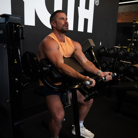 Man in orange top curls weights on arm machine in a gym. Black and yellow decor.