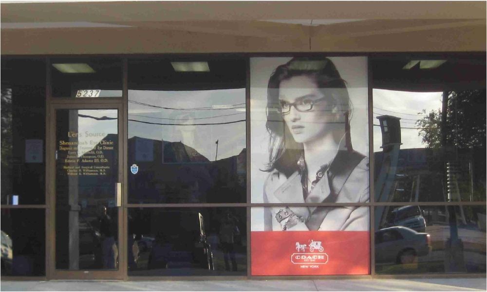 Storefront with large window display featuring a woman wearing glasses; the Coach logo is at the bottom. Reflections show the outside.