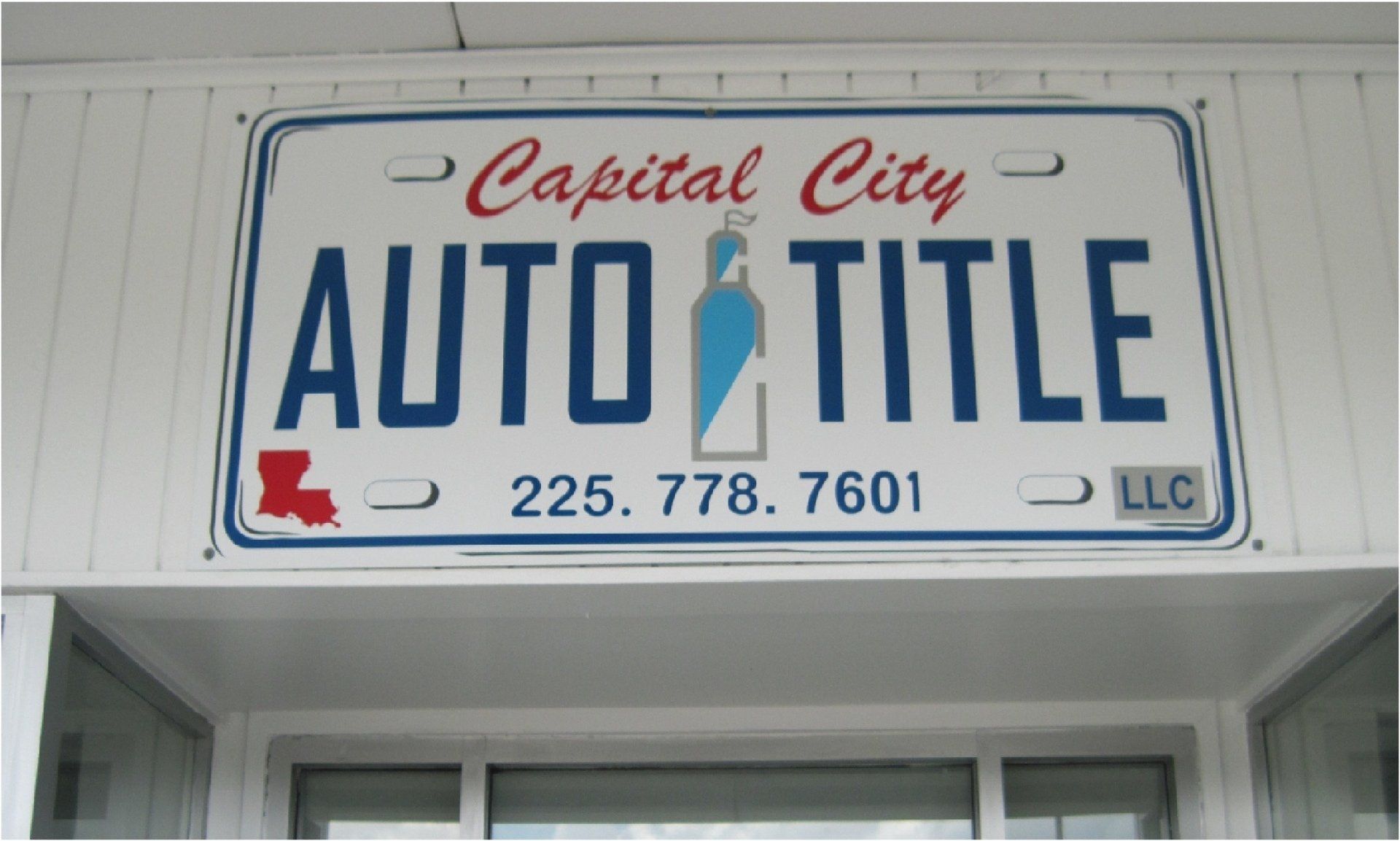 Sign for Capital City Auto Title, appearing as a license plate. It features the business name, phone number, a Louisiana state outline, and a blue, white, and gray graphic.