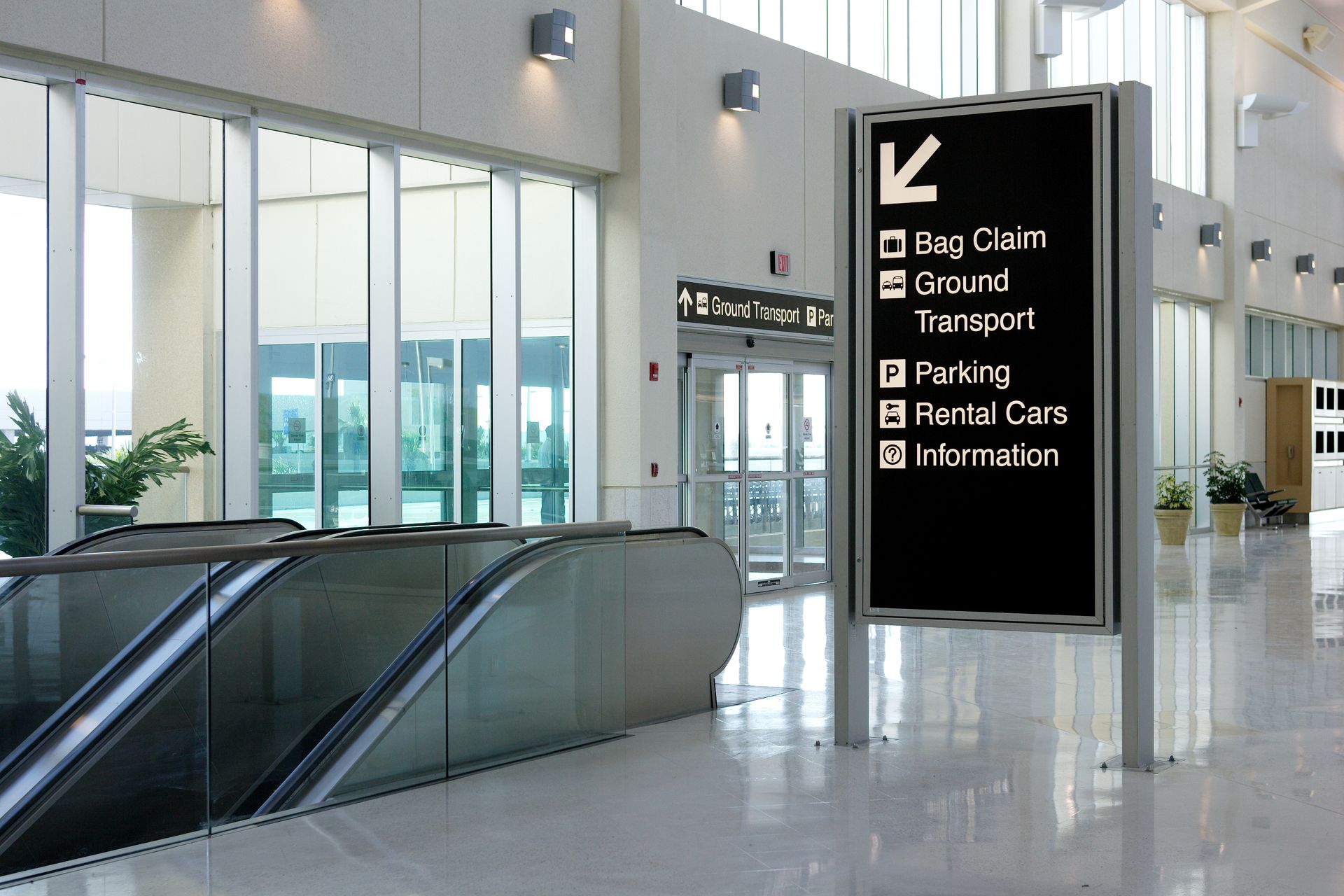 Interior airport scene with a directional sign in the center, directing towards bag claim, ground transport, parking, rental cars, and information.