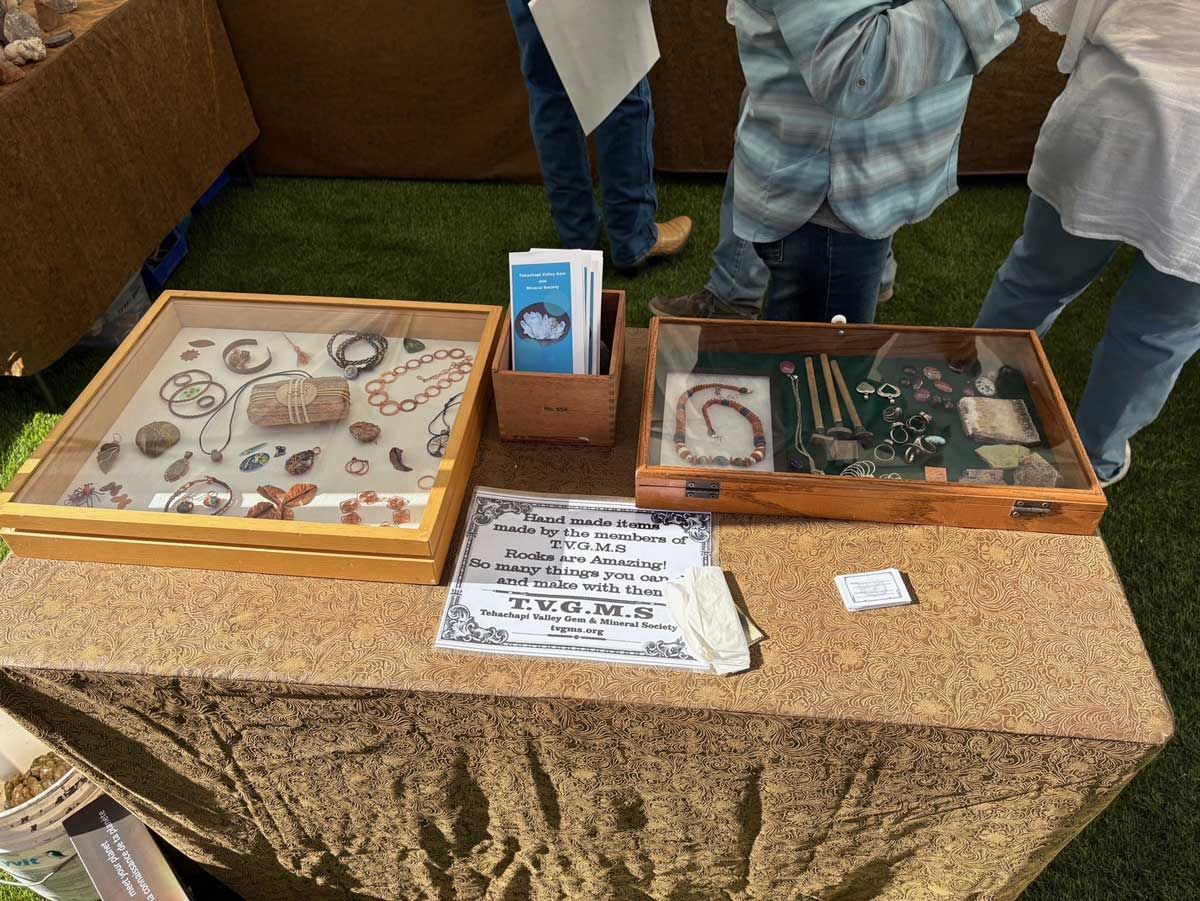 A wooden display table at an outdoor event holds two glass cases filled with jewelry and a small brochure.