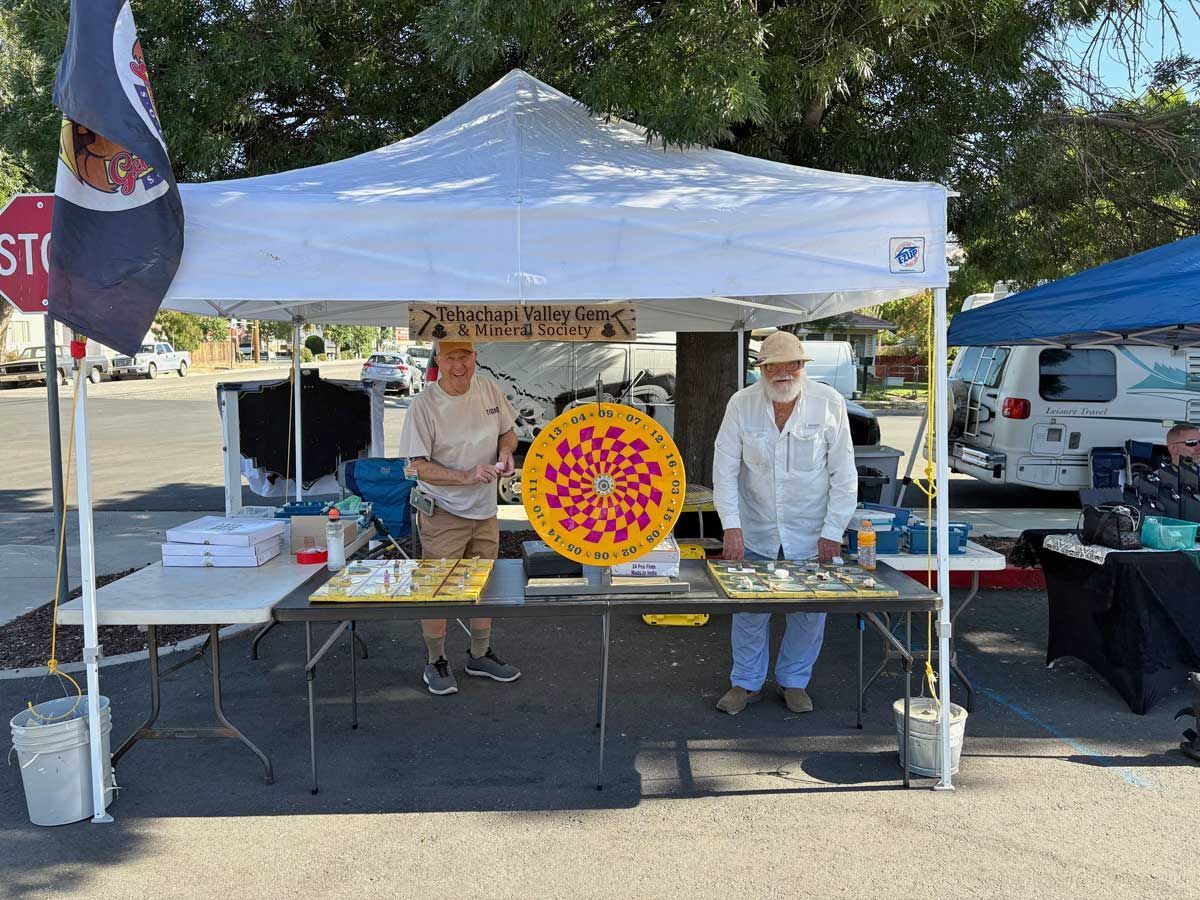 Two people stand behind a table with a spinning prize wheel and small items under a white canopy at an outdoor market.
