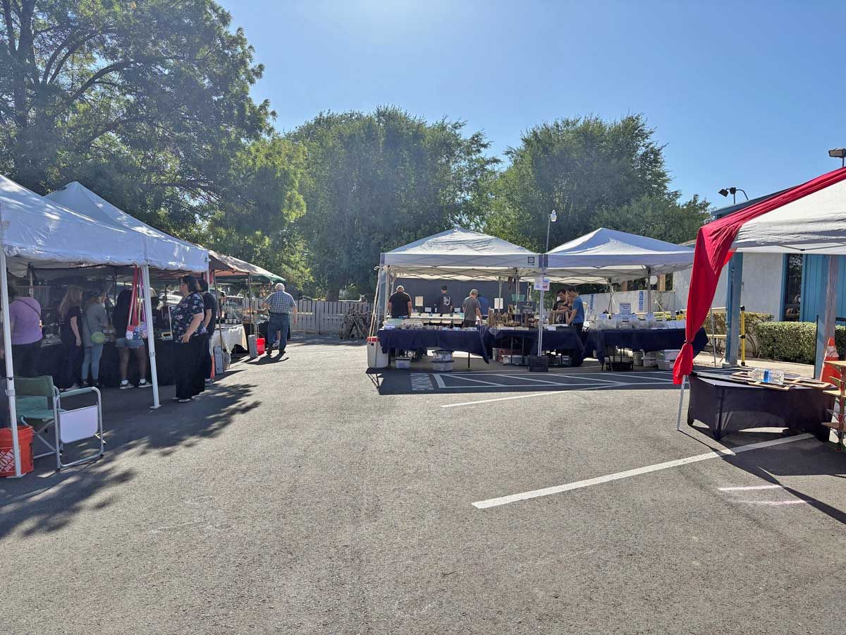 Outdoor craft market with vendor booths under white canopies on a paved parking lot on a sunny day.