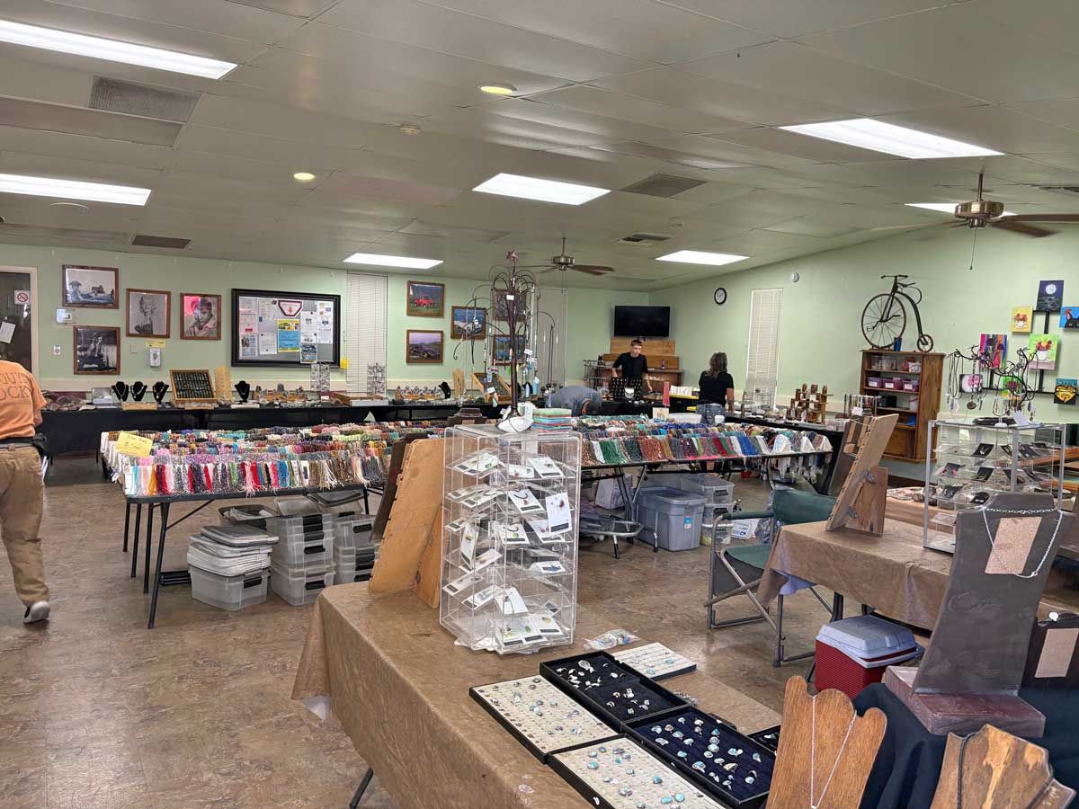 A room with long tables displaying jewelry and crafts for sale, with attendees browsing and browsing displays.
