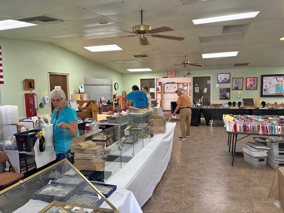 People browse displays in a bright, indoor community sale room featuring long tables covered in various goods.