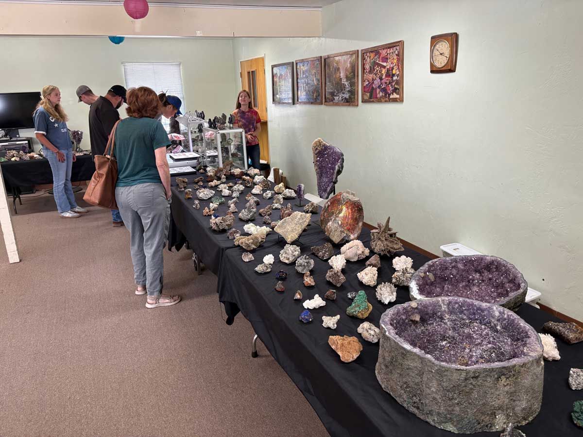 People browse a long display table filled with assorted mineral specimens and large amethyst geodes at a gem show.