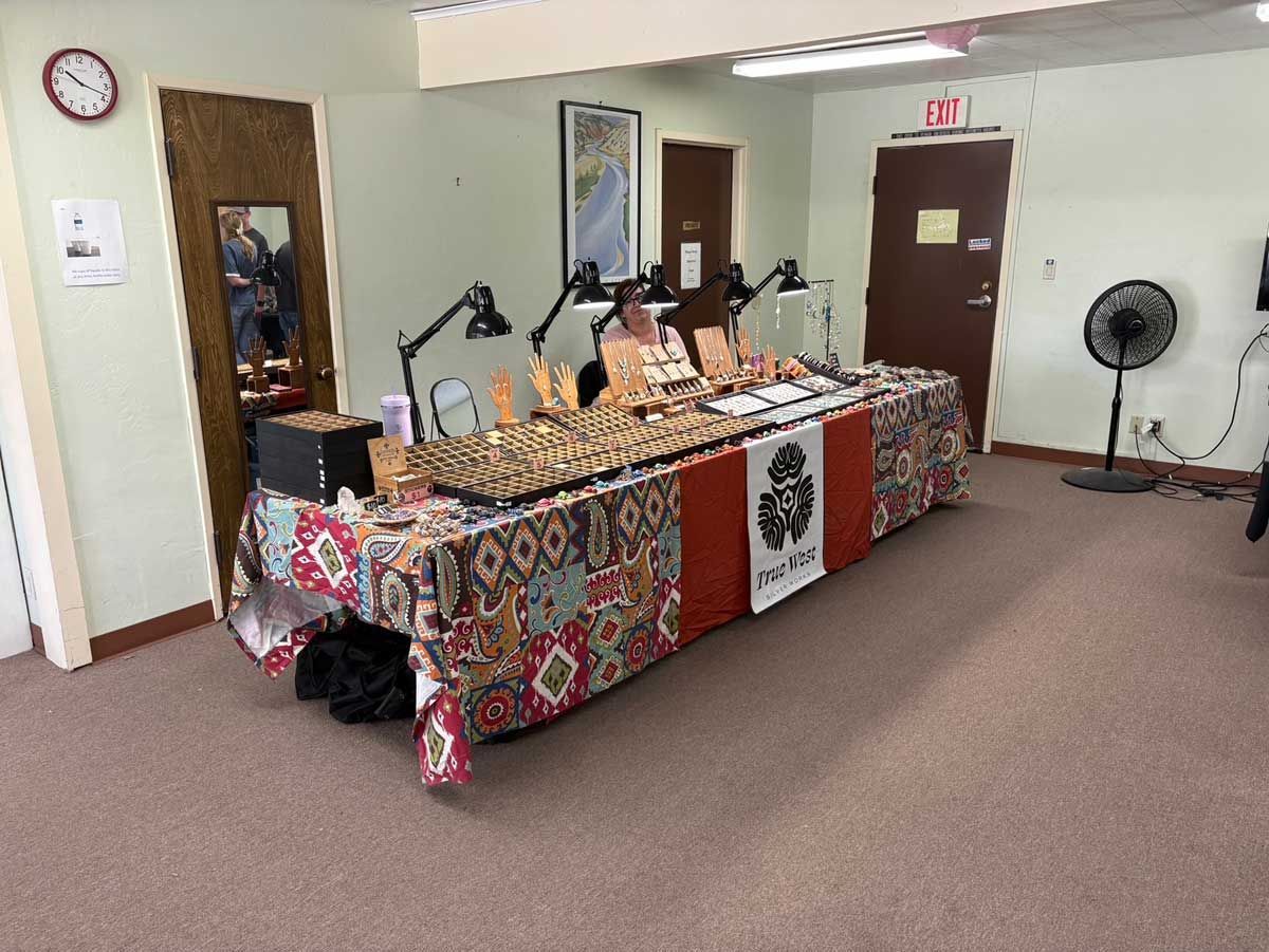 A vendor table displays jewelry in a room with a person behind it, a wall clock, and a standing floor fan.