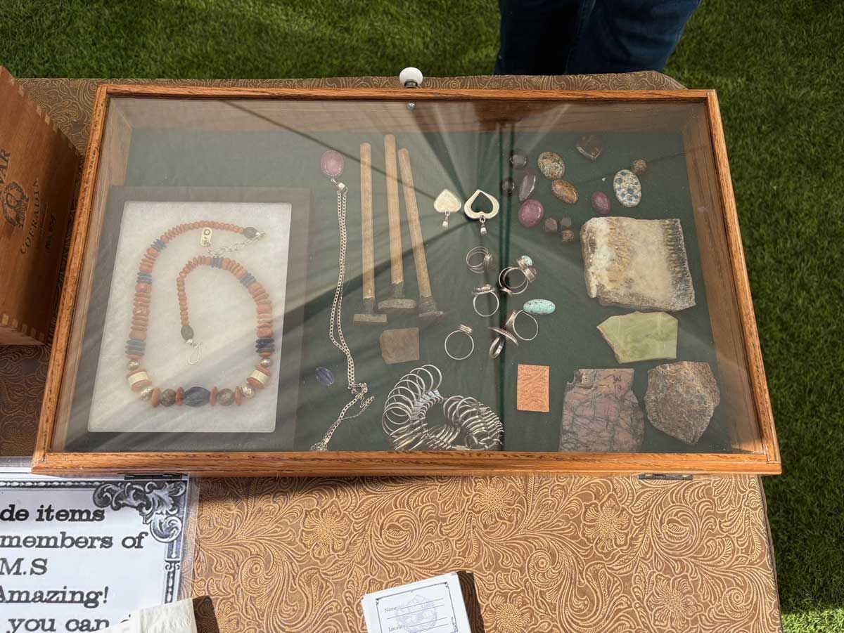 A glass-topped wooden display case on a textured table filled with assorted handmade jewelry and polished stones.