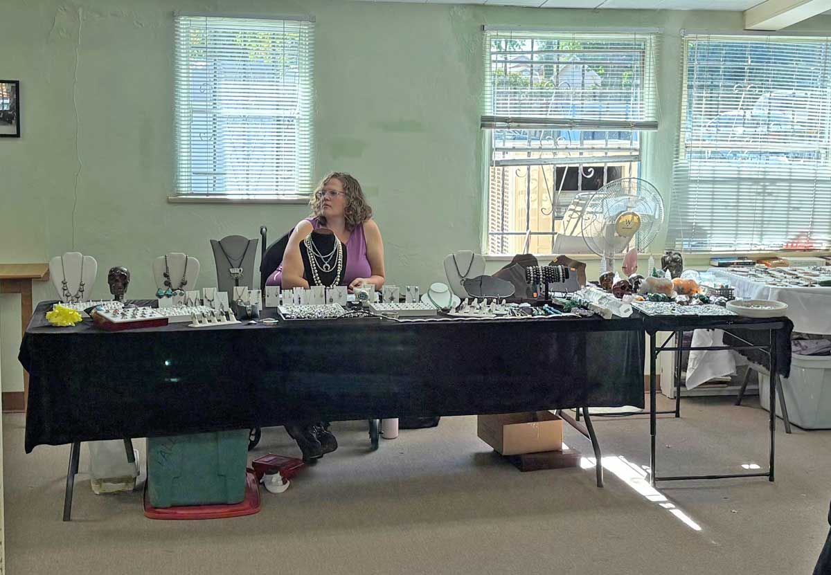 A person sitting behind a long table covered in a black cloth, displaying various jewelry items for sale in a bright room.