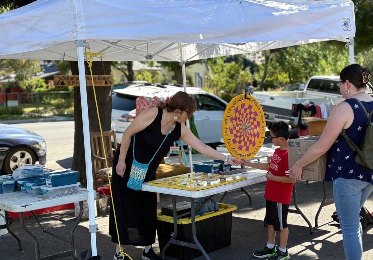 People at an outdoor booth under a white canopy, with a colorful prize wheel and tables of items.