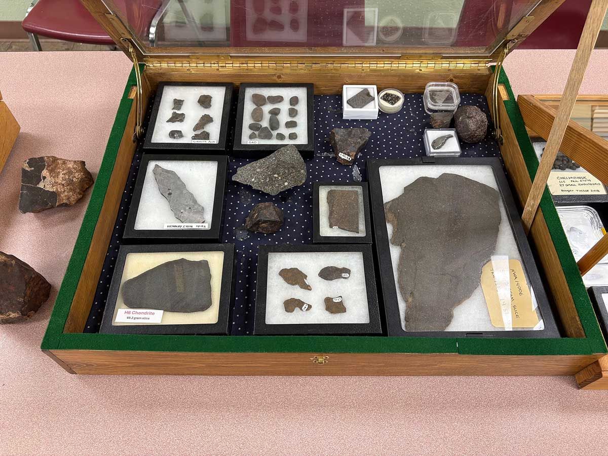Open display case with assorted mineral and fossil specimens in black-framed mounts.