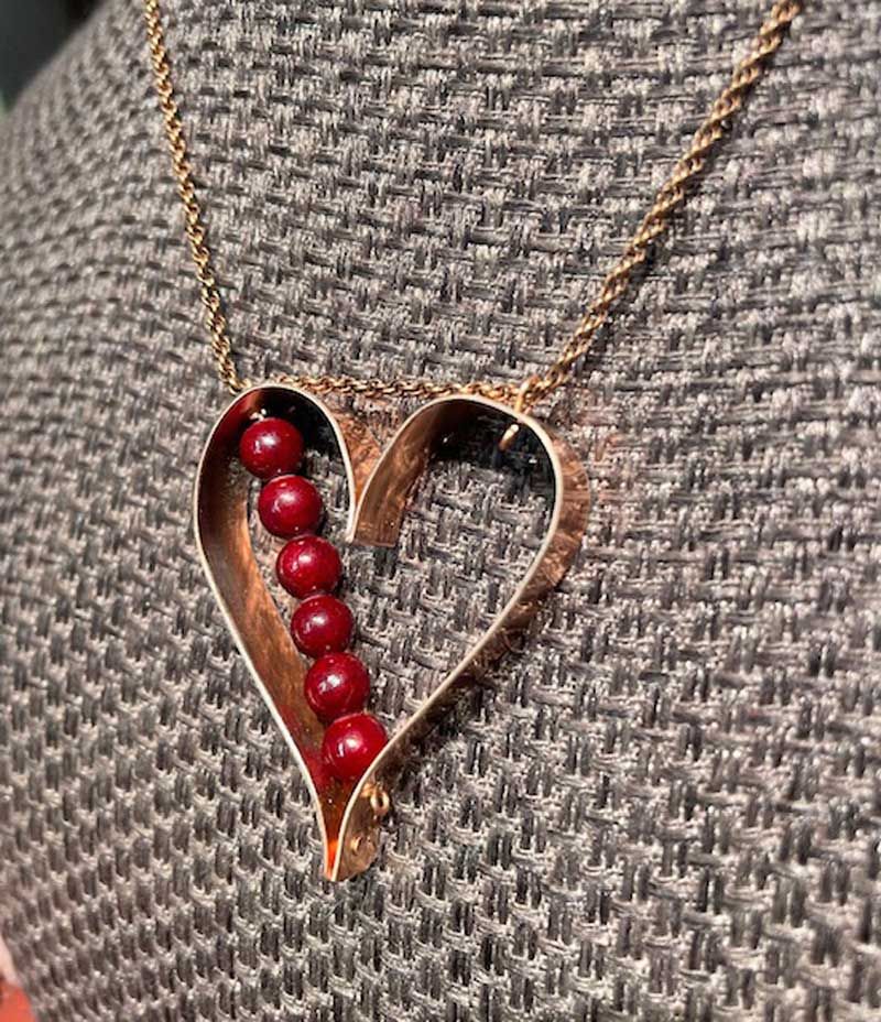 A copper-colored heart pendant on a delicate chain, featuring a vertical row of six small, deep-red beads inside the frame.