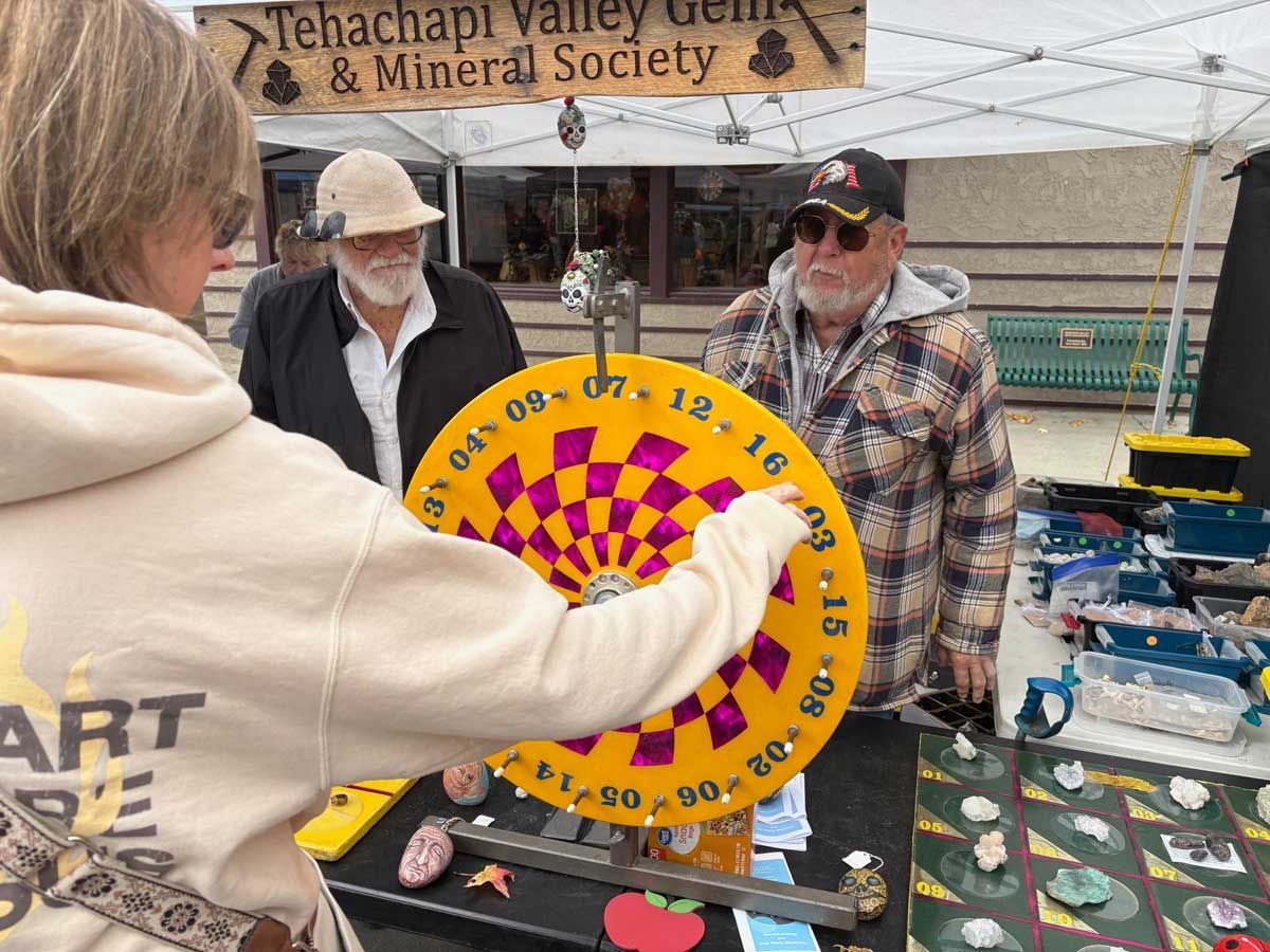 Person spins a colorful prize wheel at an outdoor market booth with two men watching.