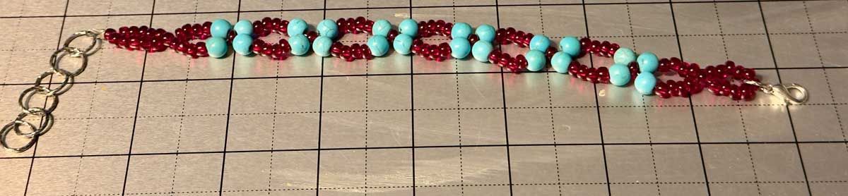 A beaded bracelet with alternating patterns of small red beads and larger turquoise spheres on a gray grid mat.