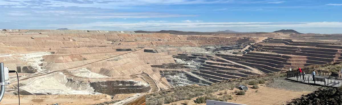 Aerial view of a large open-pit mine with terraced earth, vehicles, and dust under a blue sky