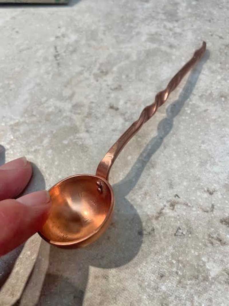 A person holds a small, hammered copper spoon with a long, decorative twisted handle against a stone surface.