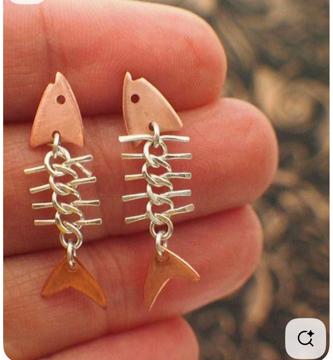 A hand holds two dangling fish-skeleton earrings made of copper heads and tails with silver chain-link spines.
