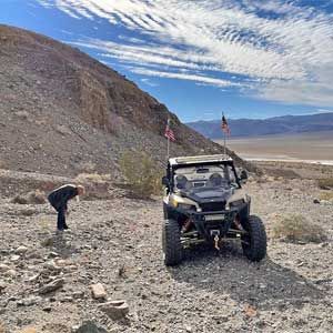 Off-road buggy on rocky desert trail, with a person bending nearby under a cloudy sky.