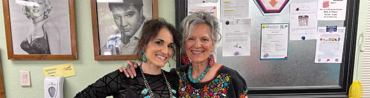 Two women smiling in front of a bulletin board with notices and a whiteboard.