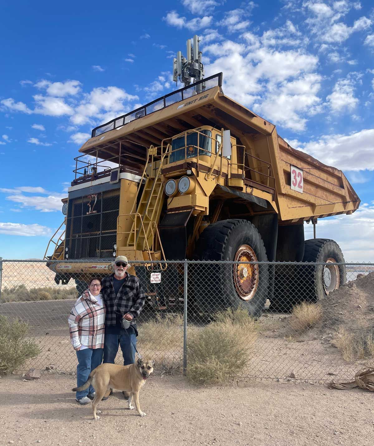 Two people and a dog stand by a giant mining truck behind a fence under a blue sky.