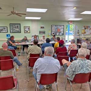 Community meeting in a bright room, with people seated in red chairs facing a panel at the front.