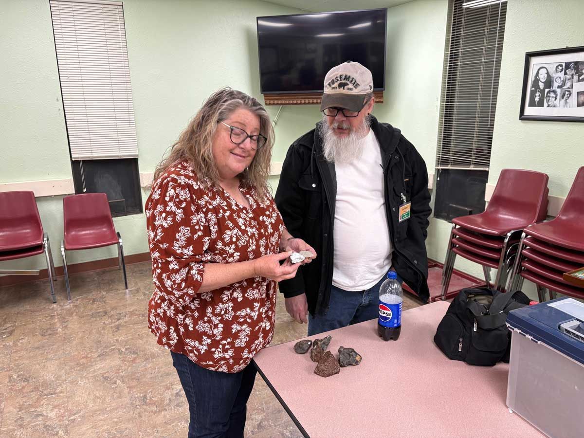 A woman and a person with a beard examine small rocks on a table in a community room with red chairs and a TV.