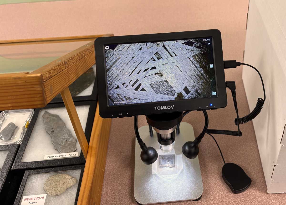 A digital microscope on a stand displays a magnified, cross-hatched pattern of a meteorite next to a display of rocks.